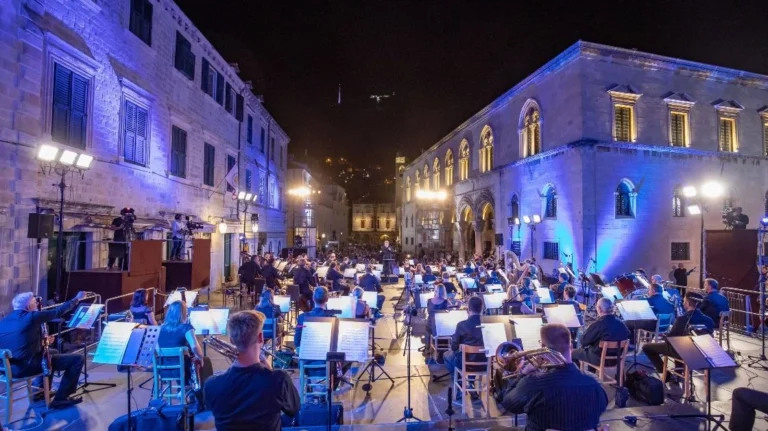 An evening open-air orchestra concert in a historic square, illuminated by blue lighting, with musicians performing under the night sky.